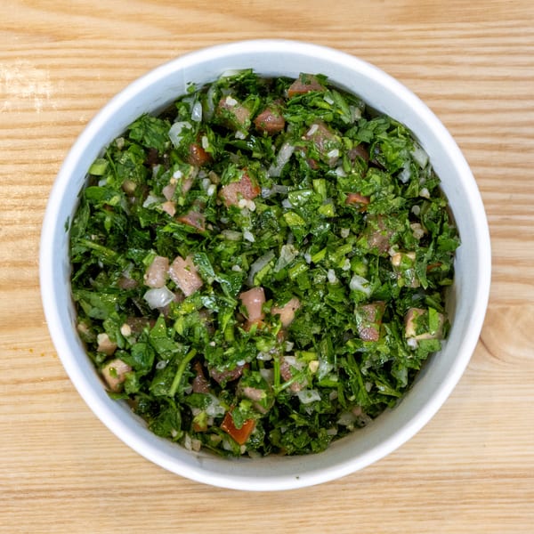 A white bowl filled with fresh green tabbouleh salad, featuring chopped parsley, tomatoes, and onions.