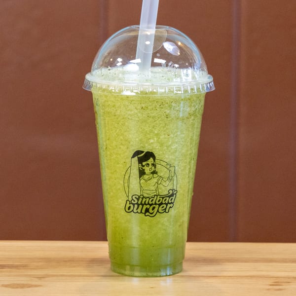 A green Sindbad Burger smoothie in a clear plastic cup with a straw on a wooden table.