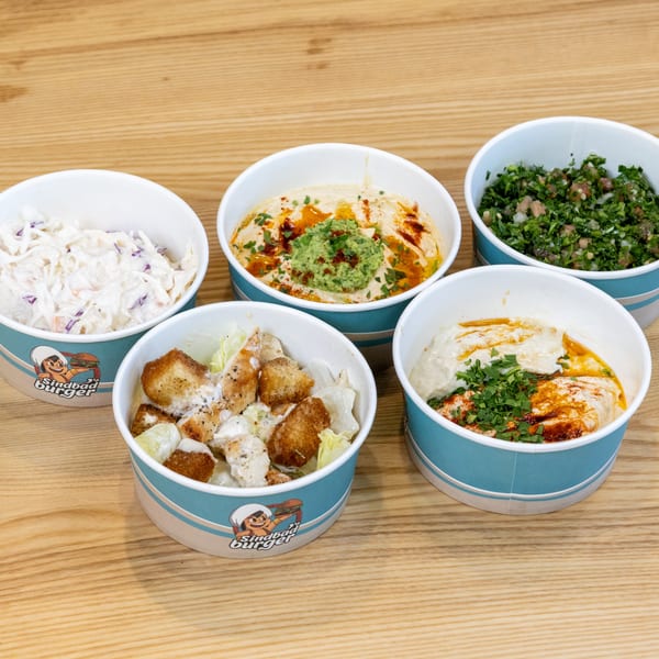 Five Sindbad Burger side dishes, including hummus, coleslaw, tabbouleh, and chicken salad.