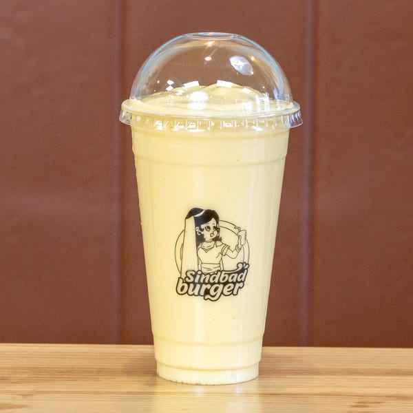 A Sindbad Burger milkshake in a clear plastic cup with a dome lid on a wooden table.