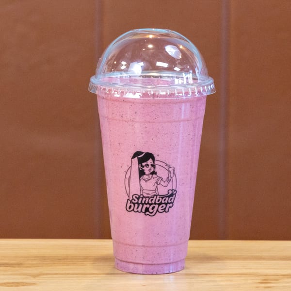 A pink Sindbad Burger smoothie in a clear plastic cup with a dome lid on a wooden table.